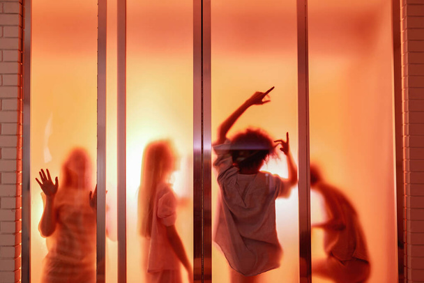 Orange silhouette of girls posing and dancing behind window of brick house during girlish sleepover at night. Girlfriends resting and having time together. Friendship. Entertainment and leisure - Zdjęcie, obraz