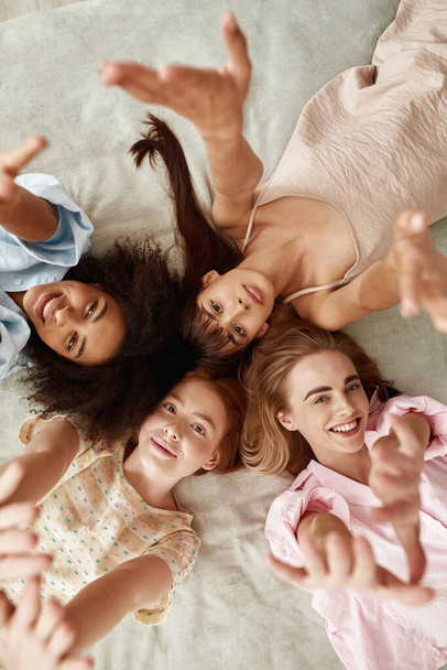 Top view of young beautiful multiracial girlfriends lying on bed and pulling hands up at girlish pajama party at home. Black and caucasian zoomer girls resting and spending time together. Friendship - Zdjęcie, obraz
