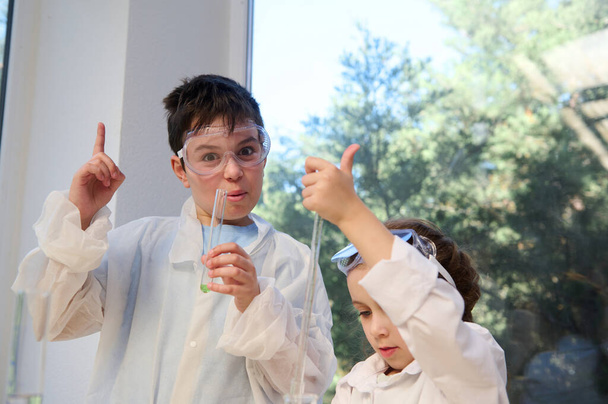 Cheerful smart kids in white lab coat and safety goggles making experiments at chemistry class. Learning chemistry science in the new semester of academic year. Back to school. Open for new knowledges - Foto, immagini