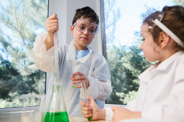 Ευρωπαϊκοί χαριτωμένοι μαθητές, αγόρια και κορίτσια μαθαίνουν επιστήμες, έρευνα και κάνουν ένα χημικό επιστημονικό πείραμα, αναλύοντας και αναμιγνύοντας υγρό σε φιάλες και δοκιμαστικούς σωλήνες στην τάξη στο σχολικό εργαστήριο - Φωτογραφία, εικόνα