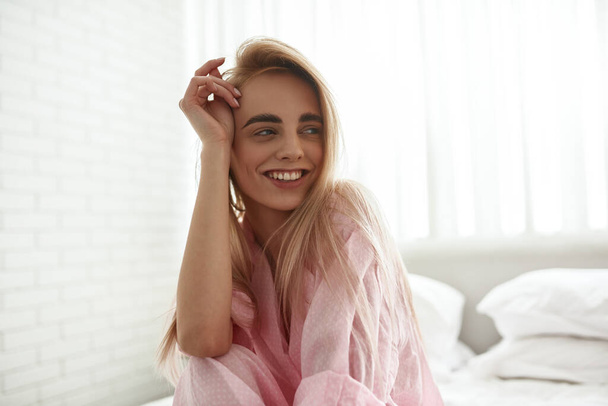 Young smiling caucasian girl sitting and looking away on bed at home in morning time. Pretty blonde teen female zoomer. Concept of domestic lifestyle. Modern bedroom in spacious apartment - Fotografie, Obrázek