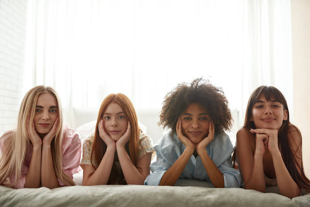 Young beautiful smiling multiethnic girlfriends lying on bed and looking at camera during girlish pajama party. Black and caucasian zoomer girls resting and spending time together. Friendship. Bedroom - Photo, image