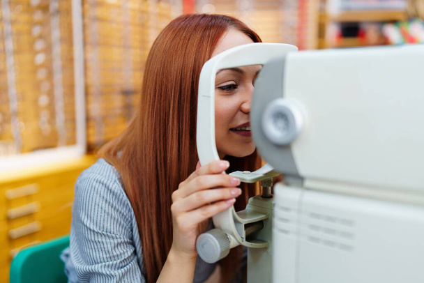 Beautiful young redhead woman is smiling during eyesight diopter examination and checkup at eye specialist clinic or optical store. - Фото, изображение