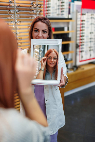 Beautiful and fashionable redhead woman choosing eyeglasses frame in modern optical store. Female seller specialist helps holding a mirror and helping her to make right decision. - Foto, Imagen