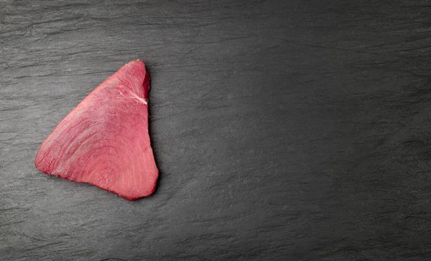 Raw tuna steak on black stone with copy space. Red sea fish fillet, fresh tuna filet, seafood sashimi, bluefin piece, akami, ahi loin, albacore meat cut on black background top view - Fotografie, Obrázek