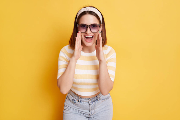 Image of positive delighted dark haired woman wearing striped t shirt and jeans posing isolated over yellow background, standing with hands near mouth and screaming, making an announce. - Foto, imagen