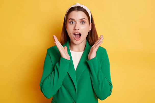 Portrait of extremely surprised woman wearing green jacket and hair band, raised arms, keeps mouth widely opened, being very shocked, posing isolated over yellow background. - Photo, image