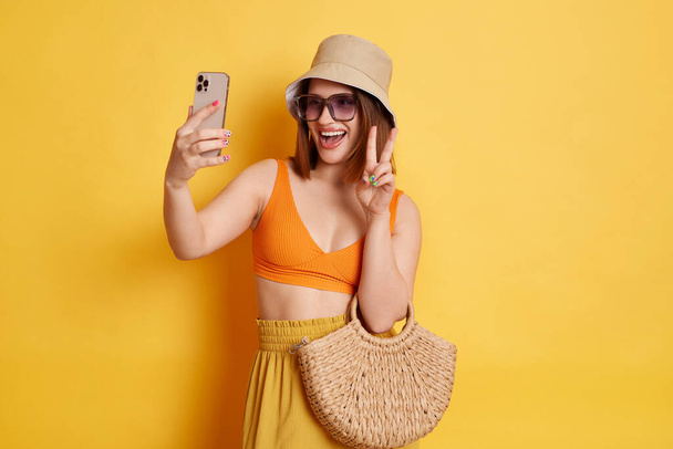 Indoor shot of positive woman wearing orange top and panama posing isolated over yellow background, having rest at resort, holding cell phone, having video call, showing v sign. - Фото, зображення