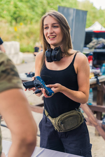 Smiling white woman with safety headphones around neck buying handgun from man. Buying and selling firearms topic. Outdoor vertical shot. High quality photo - Foto, Imagem