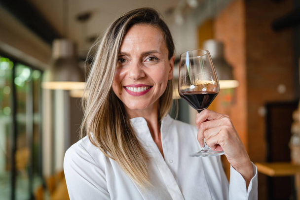 Одна женщина среднего возраста белая женщина, стоящая дома в столовой или ресторане счастливая улыбка уверенно с бокалом красного вина носить белую рубашку талии вверх копия пространства фронтальный вид портрет - Фото, изображение