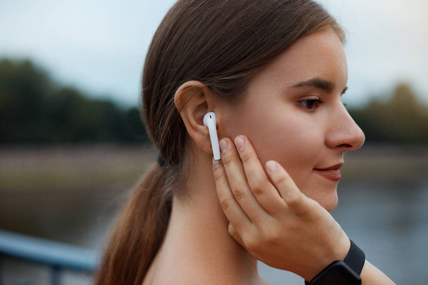 Close up portrait of a lovely young fitness girl listening to music through wireless earphones outdoors. - Foto, immagini