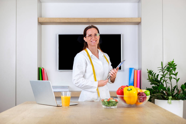 Portrait of a smiling young woman doctor nutritionist standing holding weighing scale and tape measure in bright modern office room - Photo, image
