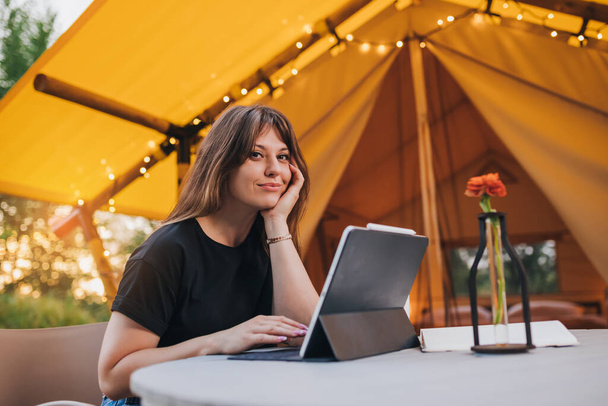 Happy Woman freelancer using a laptop on a cozy glamping tent in a sunny day. Luxury camping tent for outdoor summer holiday and vacation. Lifestyle concept - 写真・画像