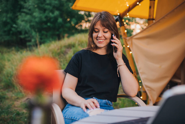 Happy Woman freelancer talking phone and using laptop on cozy glamping tent in a sunny day. Luxury camping tent for outdoor summer holiday and vacation. Lifestyle concept - Foto, Imagem