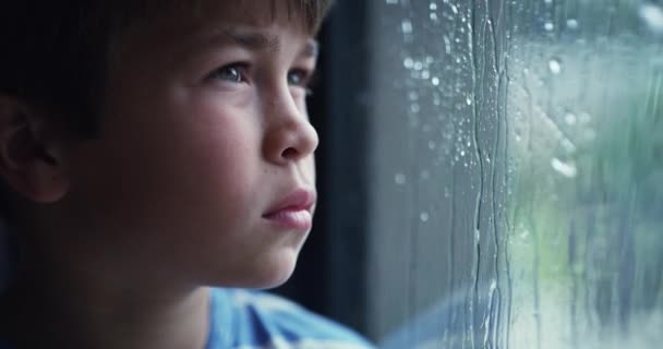 A sad little boy depressed by bad weather, sitting inside watching rain through a window. Disappointed child unhappy, bored, lonely and frustrated by failed plans. Kid stuck indoors due to a storm. - Filmati, video