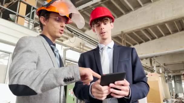 Dva inženýři diskutují o práci pomocí tabletu, jsou dobře oblečeni a nosí ochranné přilby. - Záběry, video