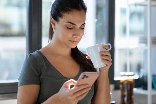 Shot of beautiful young woman using her mobile phone while drinking a cup of coffee in the kitchen at home. - Photo, image