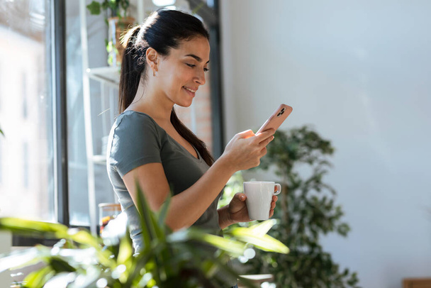 Shot of beautiful young woman using her mobile phone while drinking a cup of coffee in the kitchen at home. - Foto, Imagen