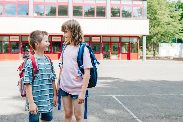 Хлопчик і дівчинка, брат і сестра стоять з рюкзаками на спині перед будівлею школи і розмовляють. Діти повертаються до школи після канікул
. - Фото, зображення