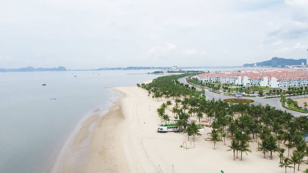 Aerial view tropical beach in Ha Long Bay with Tuan Chau island and hotel resort in background. Tourist destination in Vietnam with white sandy shoreline, palm trees and recreational activities - 写真・画像