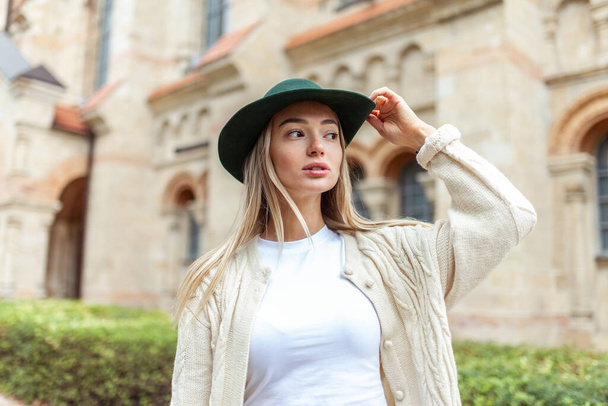Вулична мода, спосіб життя. Модна жінка блондинка в стильному капелюсі позує на тлі старої міської архітектури
 - Фото, зображення