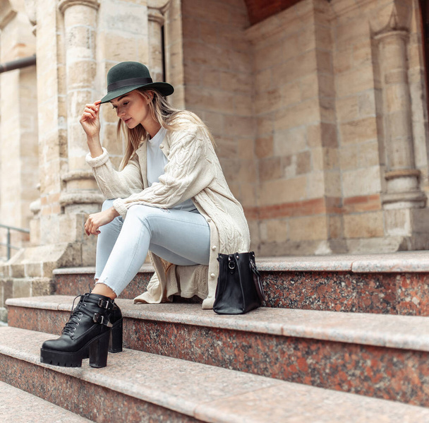 Young fashion woman in trendy stylish clothes sits on steps in urban architecture. Lifestyle, street fashion - Zdjęcie, obraz