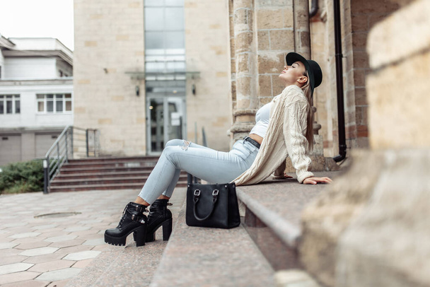 Young fashion woman in trendy stylish clothes sits on steps in urban architecture. Lifestyle, street fashion - Foto, Imagem