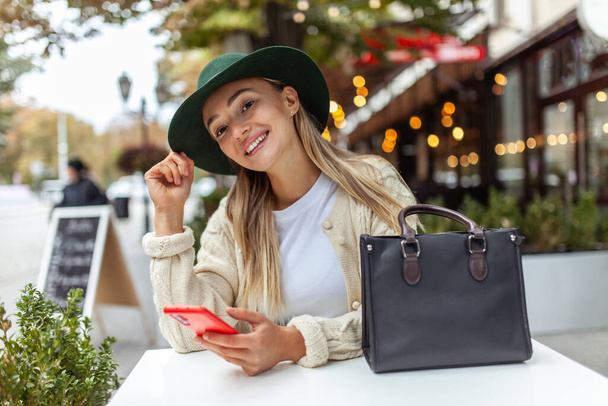 Cute fashion woman smiling and using smartphone while sitting at a table in an outdoor cafe. Lifestyle - Valokuva, kuva