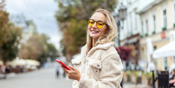 Portrait of a cute smiling fashion woman in yellow sunglasses, using smartphone in the city. Lifestyle - 写真・画像