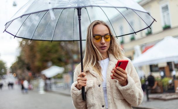 Cute fashion blonde woman in yellow sunglasses with transparent umbrella using phone in the city. Lifestyle - Fotoğraf, Görsel