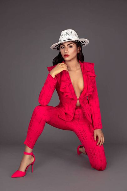 Portrait of a beautiful young white woman with wavy black hair and beautiful makeup posing by herself inside a studio with a grey background wearing a red business suit with high heels and a white hat. - Foto, Bild
