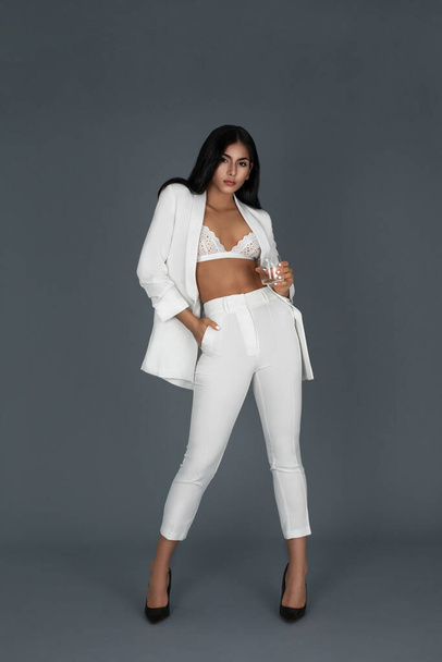 Portrait of a sexy young white woman with wavy black hair and beautiful makeup posing by herself inside a studio with a grey background wearing a white suit with black high heels and a white bra. - Photo, image