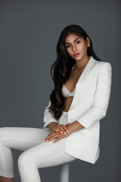 Portrait of an innocent white woman with wavy black hair and beautiful makeup posing by herself inside a studio with a grey background wearing a white suit with black high heels and white bra. - Φωτογραφία, εικόνα