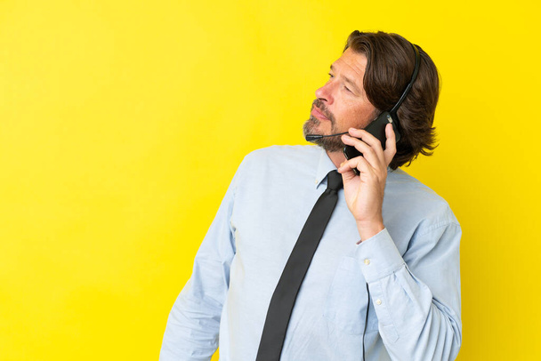 Tele pazarlamacı Hollandalı adam sarı arka planda izole edilmiş bir kulaklıkla çalışıyor cep telefonuyla sohbet ediyor. - Fotoğraf, Görsel