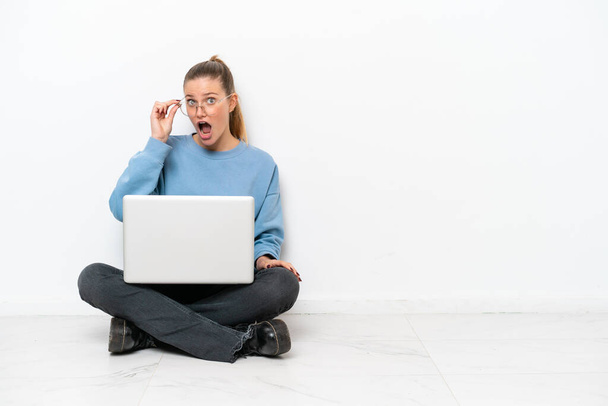 Jonge vrouw met een laptop zittend op de vloer met bril en verrast - Foto, afbeelding