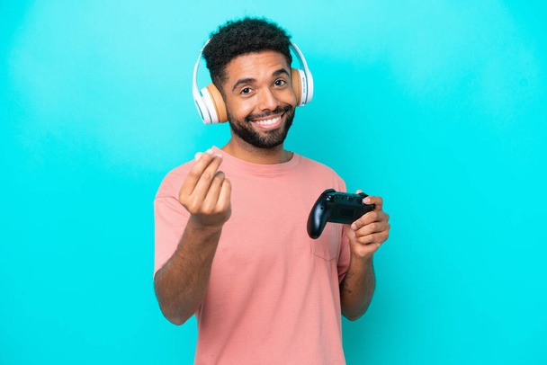 Young brazilian man playing with a video game controller isolated on blue background making money gesture - Foto, imagen