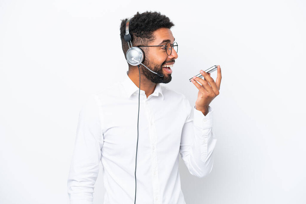 Telemarketer Brazilian man working with a headset isolated on white background keeping a conversation with the mobile phone - Foto, afbeelding