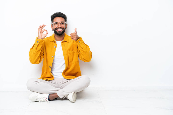 Young Brazilian man sitting on the floor isolated on white background showing ok sign and thumb up gesture - Φωτογραφία, εικόνα