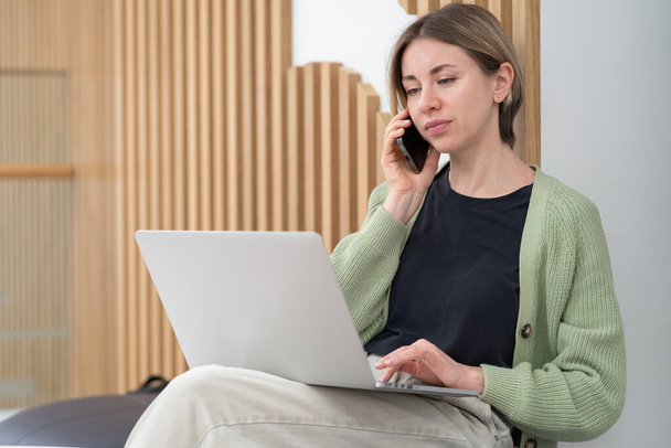 Serious woman holding smartphone discussing project details while working online on laptop in coworking space, talking with client via cellphone, businesswoman with mobile phone looking at pc screen - Foto, Imagem