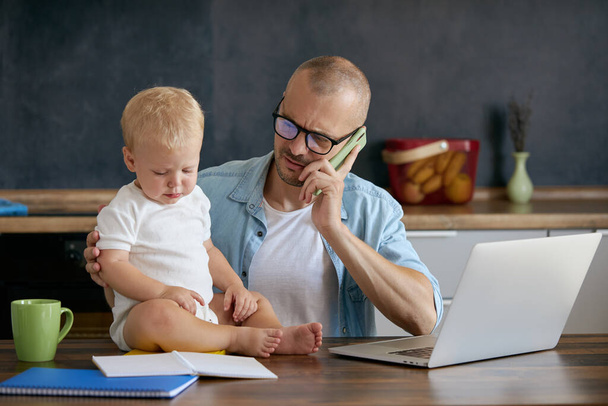 Young father freelancer working on laptop remotely from home office with baby in his arms. Concept of resilience while working from home with kids during quarantine - Foto, Bild