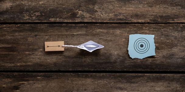 Origami boat pulling wooden peg with arrow drawn on it towards a blue paper with target sign. Conceptual image of vision and clear goals. Over rustic wooden background.  - Photo, image