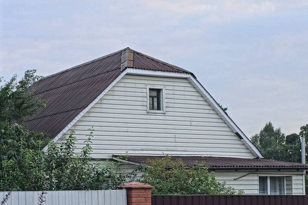 white attic of a rural house with a small window under a gray slate roof on the street against the sky - Photo, image