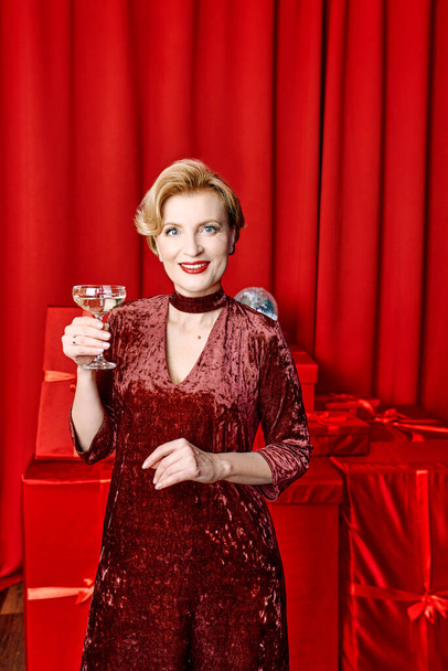 ώριμη κομψή κομψή γυναίκα με ποτήρι αφρώδους οίνου με δώρα σε κόκκινο φόντο. Κόμμα, μόδα, γιορτή, έννοια αντι ηλικία  - Φωτογραφία, εικόνα