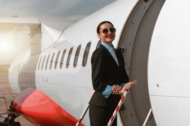 Woman flight attendant in sunglasses standing on airplane stairs at airport. Blurred background - Foto, Imagen