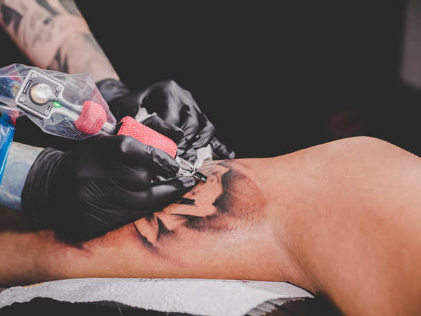 Close up view of tattoo artist demonstrates the process of getting black skull tattoo with paint. Concentrated female coloring contour ink on leg with gun. Neon light. - Foto, imagen