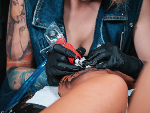 Close-up beeld van tatoeëerder toont het proces van het verkrijgen van zwarte schedel tatoeage met verf. Geconcentreerd vrouwelijke kleuren contour inkt op been met pistool. Neon licht. - Foto, afbeelding