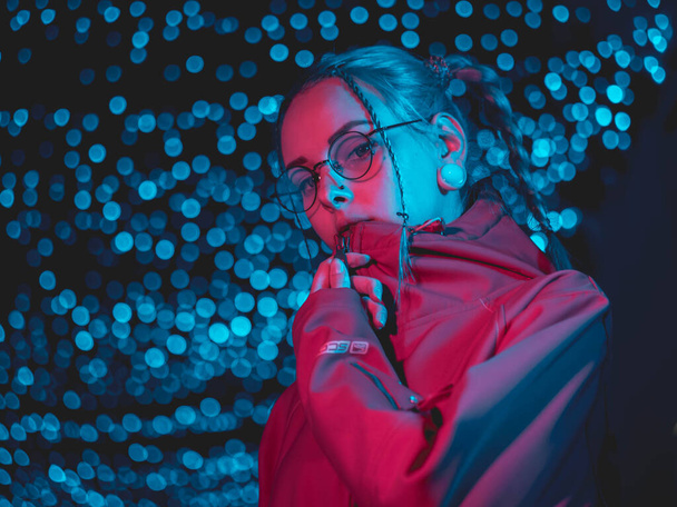 Young pretty girl with unusual hairstyle near glowing pink and blue neon lights of the city at night. Dyed blue hair in braids. Serious hipster teenager in glasses and beautiful lenses. - Fotoğraf, Görsel