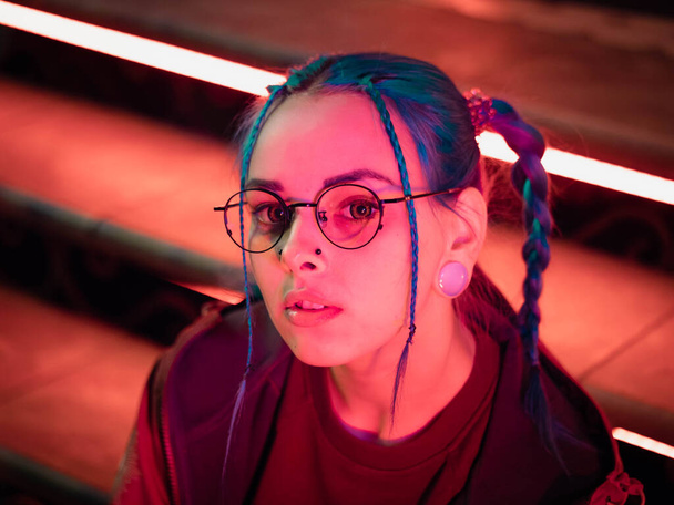 Young pretty girl with unusual hairstyle sitting on stairs which glowing red neon lights. City at night. Dyed blue hair in braids. Happy hipster teenager in glasses. - Photo, image
