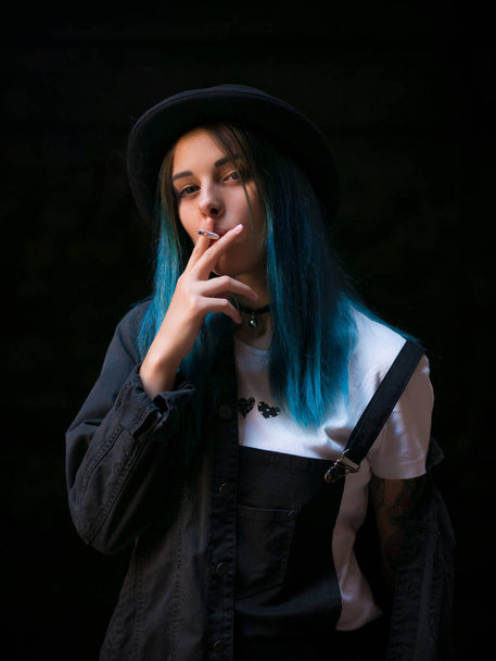 Emo girl smoking cigarette.Street punk or hipster woman with blue colorful dyed hair, hat, piercing,lenses,ears tunnels and unusual hairstyle stands in backyard. beautiful smoke. - Foto, immagini