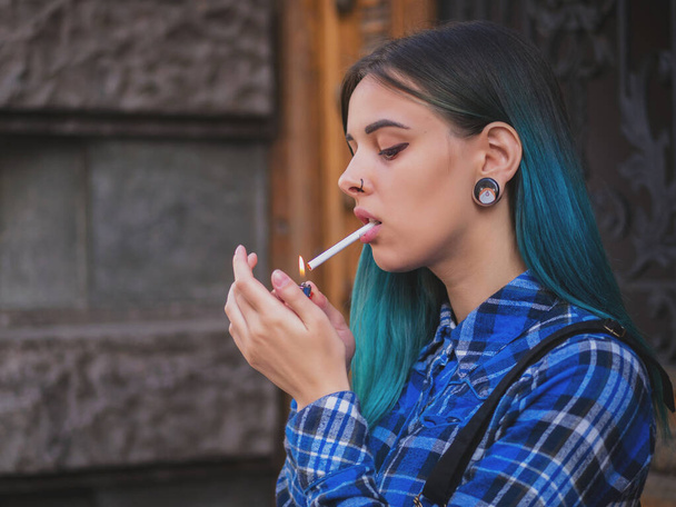 Young punk girl smoking cigarette by the wooden door.Hipster with blue dyed hair. Woman with piercing in nose, violet lenses, ears tunnels and unusual hairstyle. - Фото, зображення